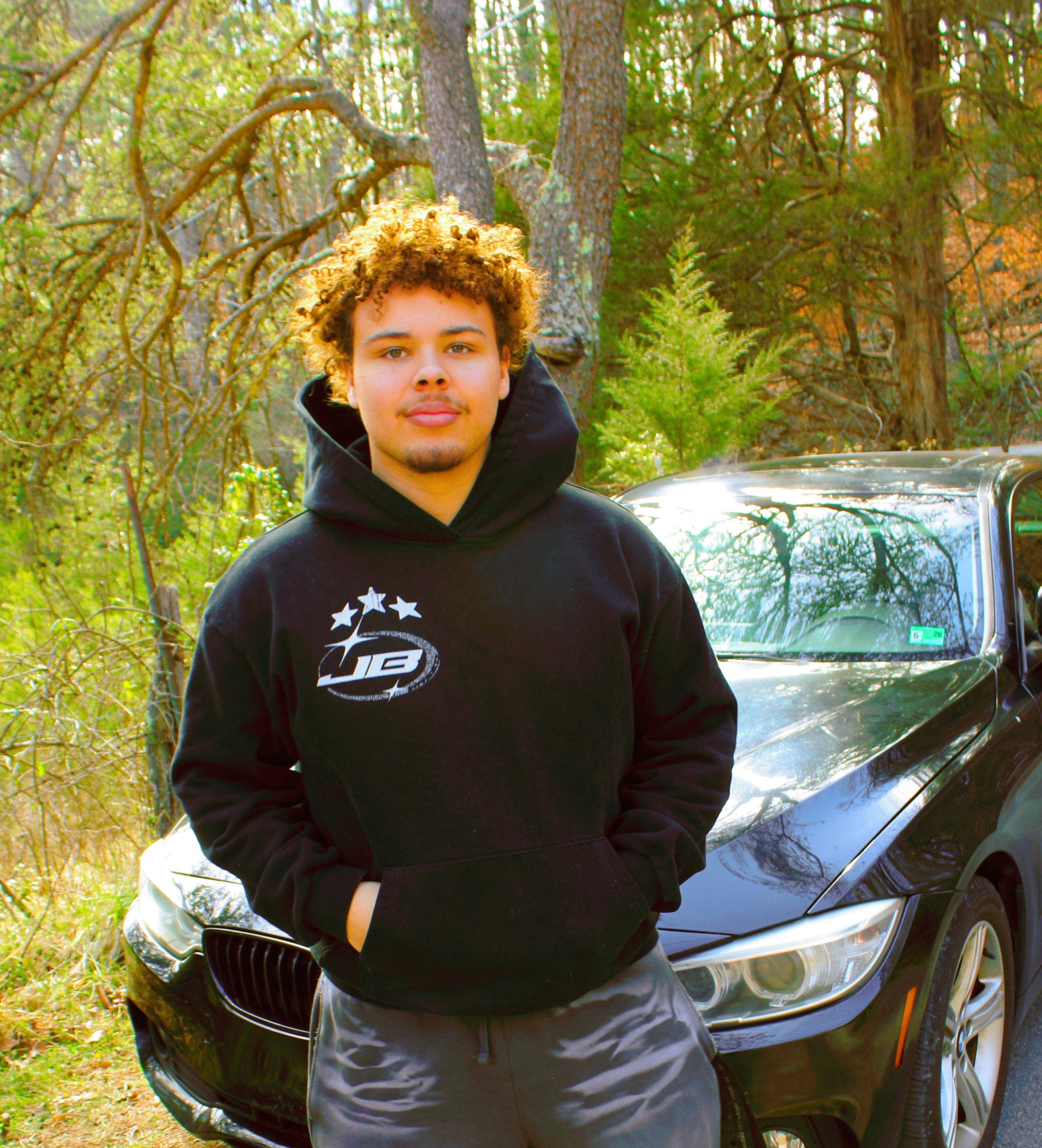 Person wearing a black hoodie and gray pants standing next to a black car in a wooded area.